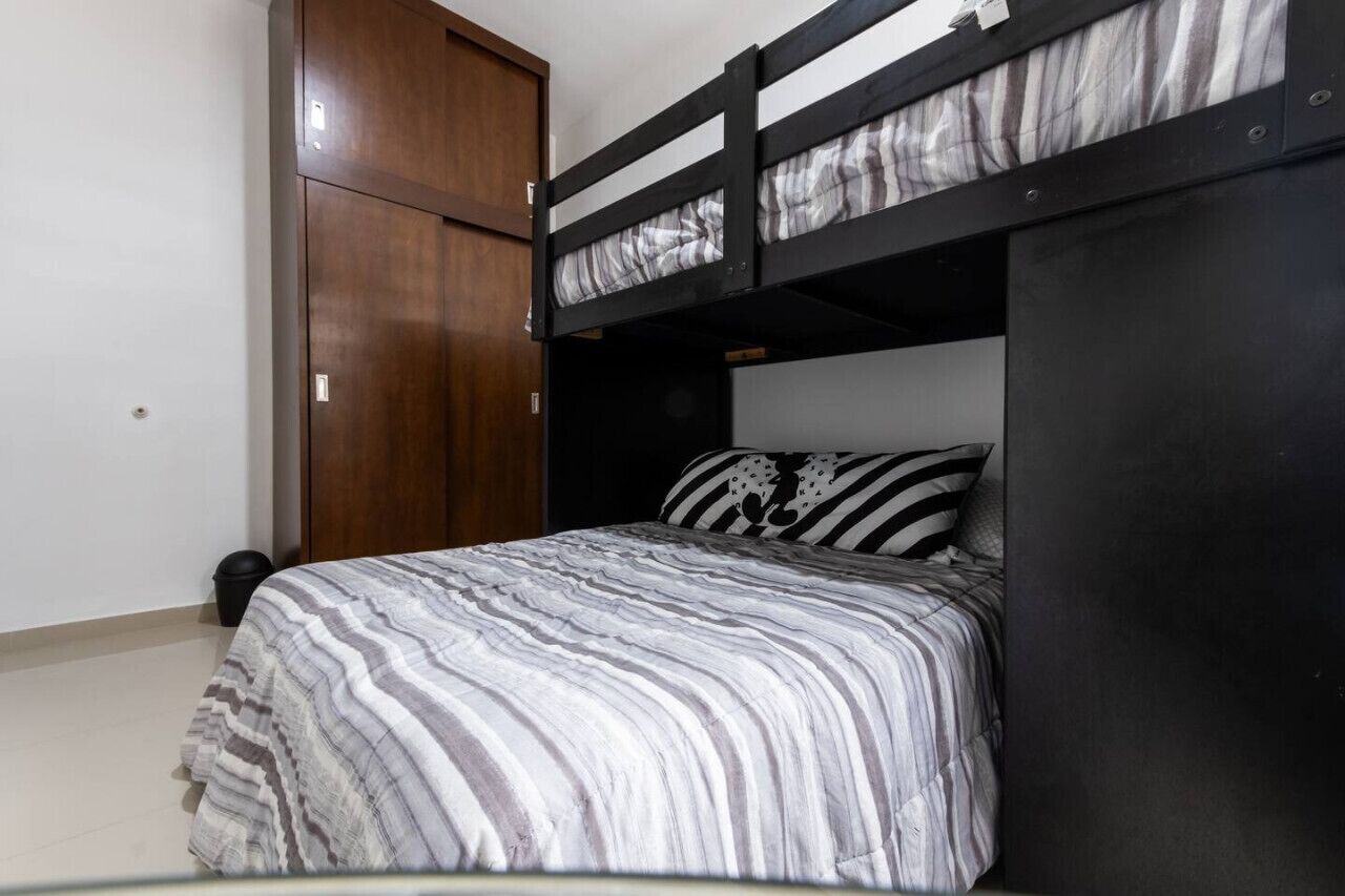 Un lit superposé avec une literie à rayures, une armoire en bois et une petite poubelle.