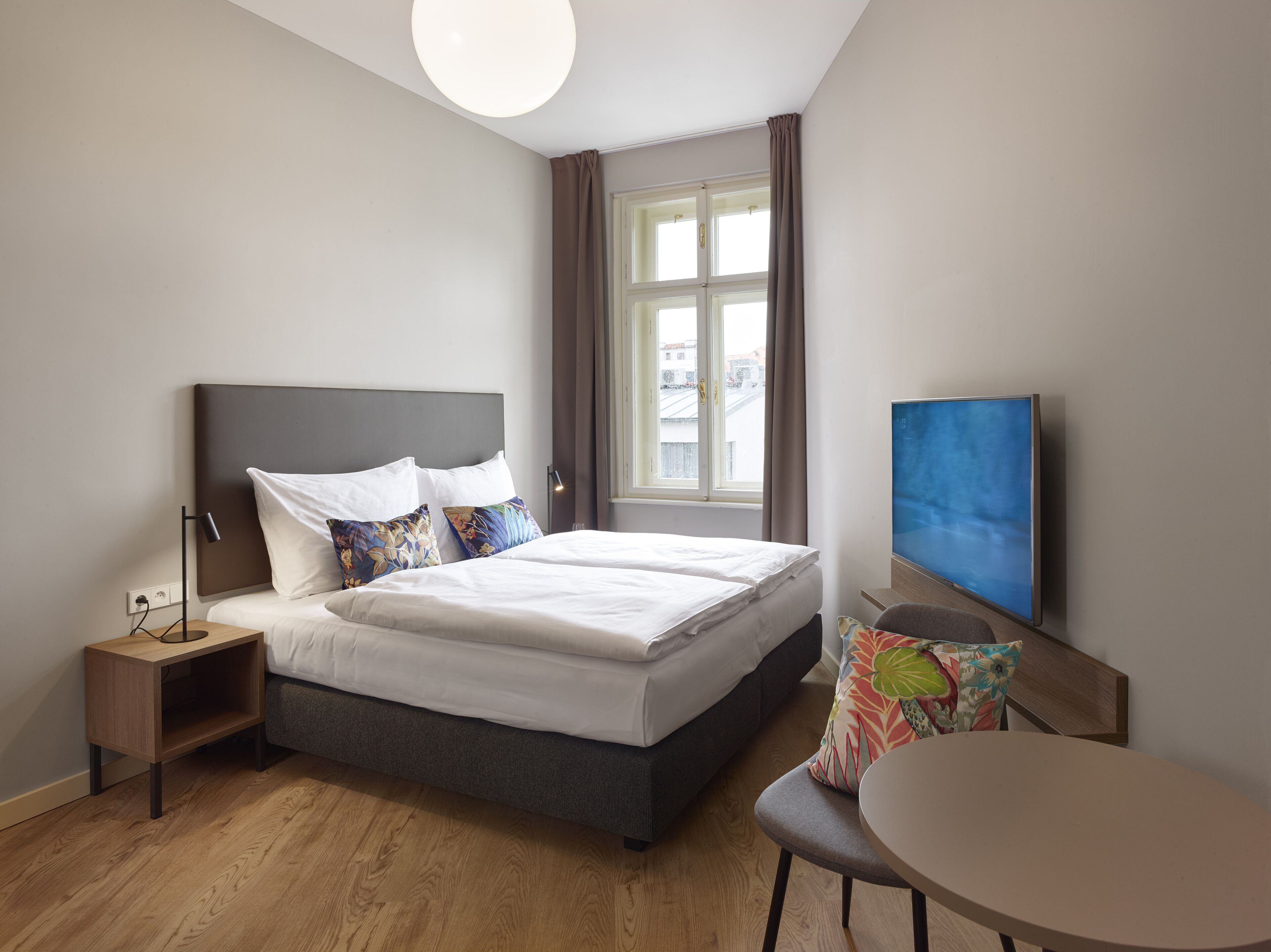 A modern hotel room with a large bed, bedside tables, a desk, a chair, a television, and a window with curtains.