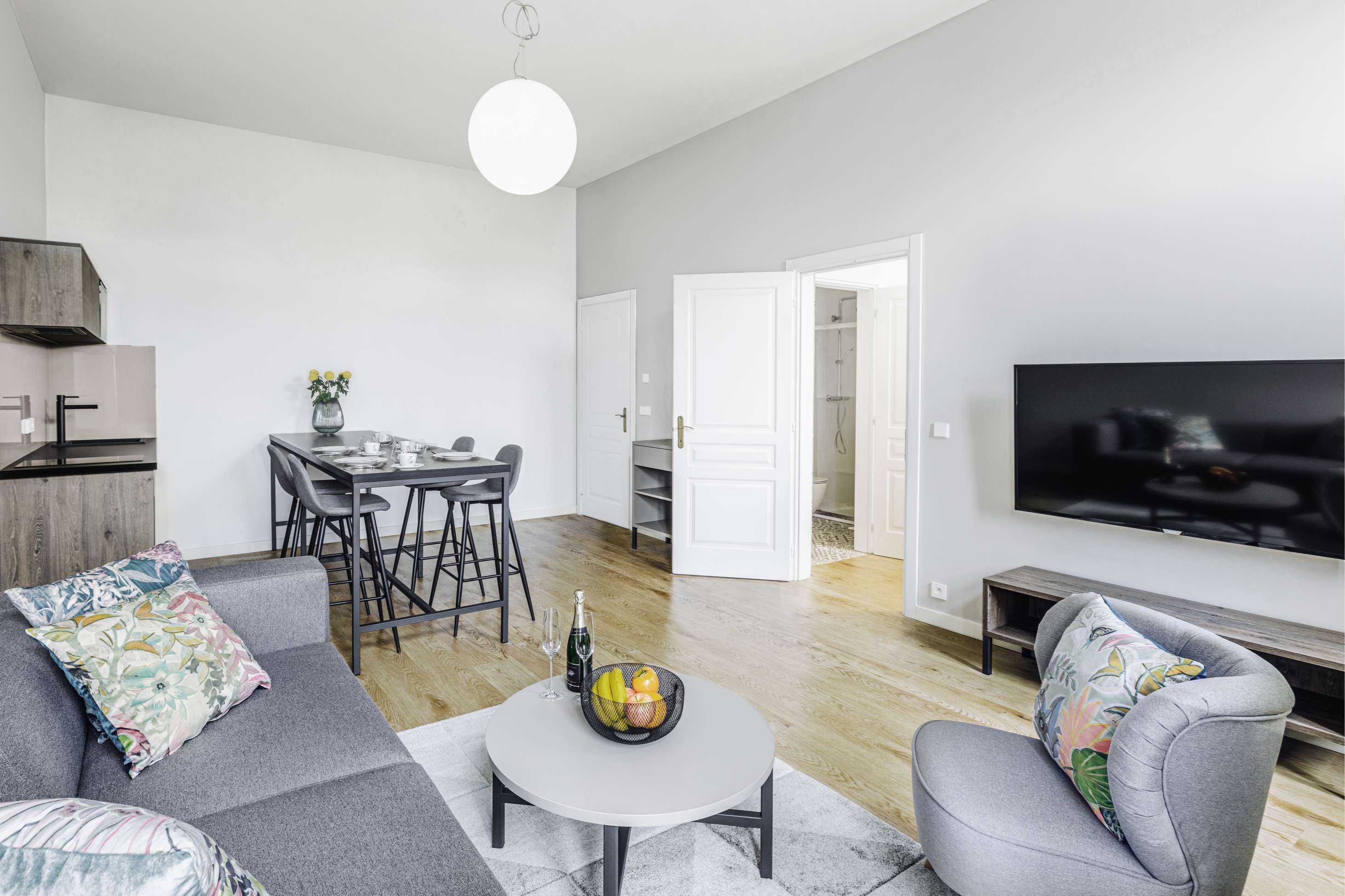 A modern living room with a grey sofa, a round coffee table with a fruit bowl, a dining area with a white table and chairs, and a TV mounted on the wall.