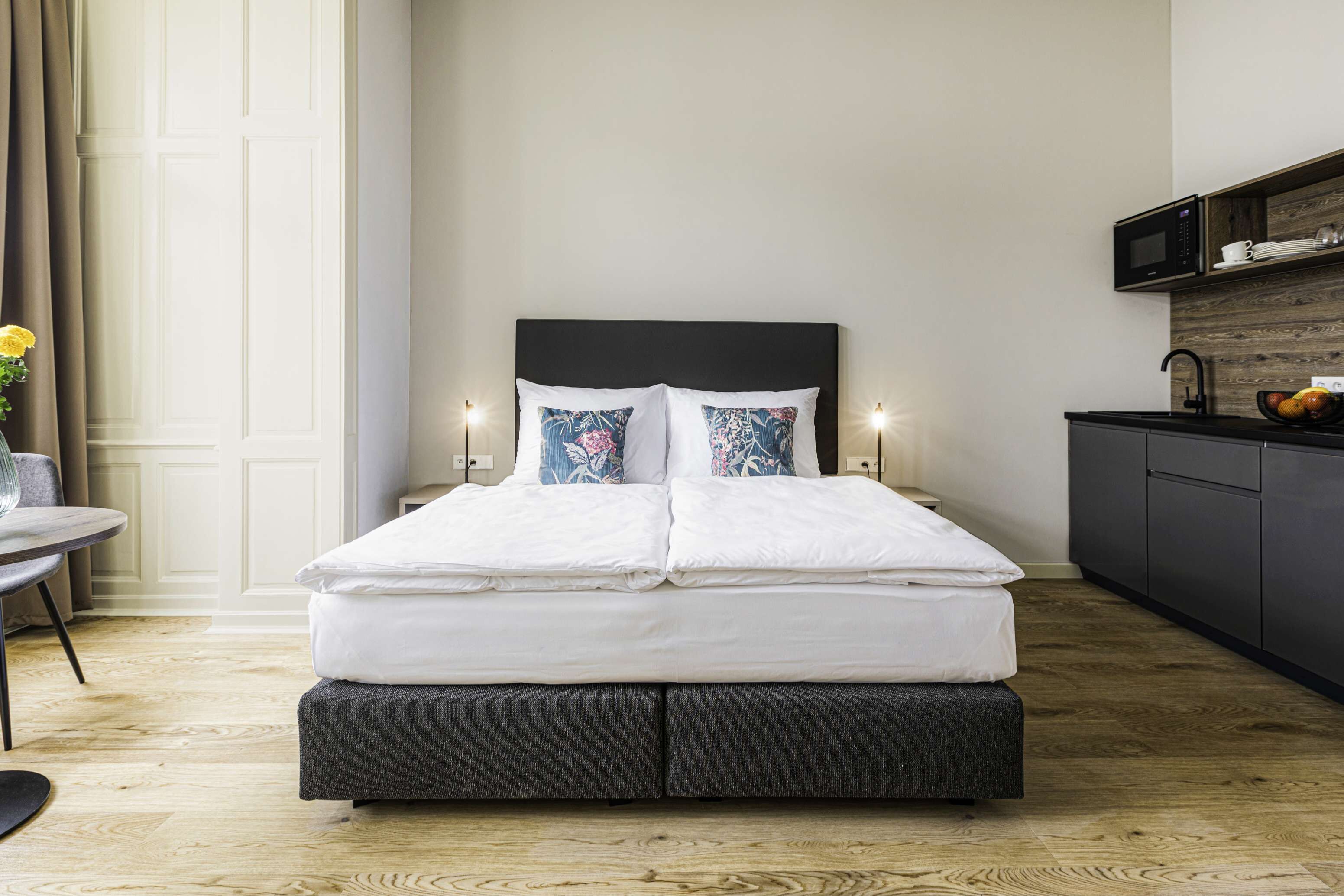Une chambre moderne avec un lit, des lampes de chevet, une petite table avec un vase et une vue sur un coin cuisine.
