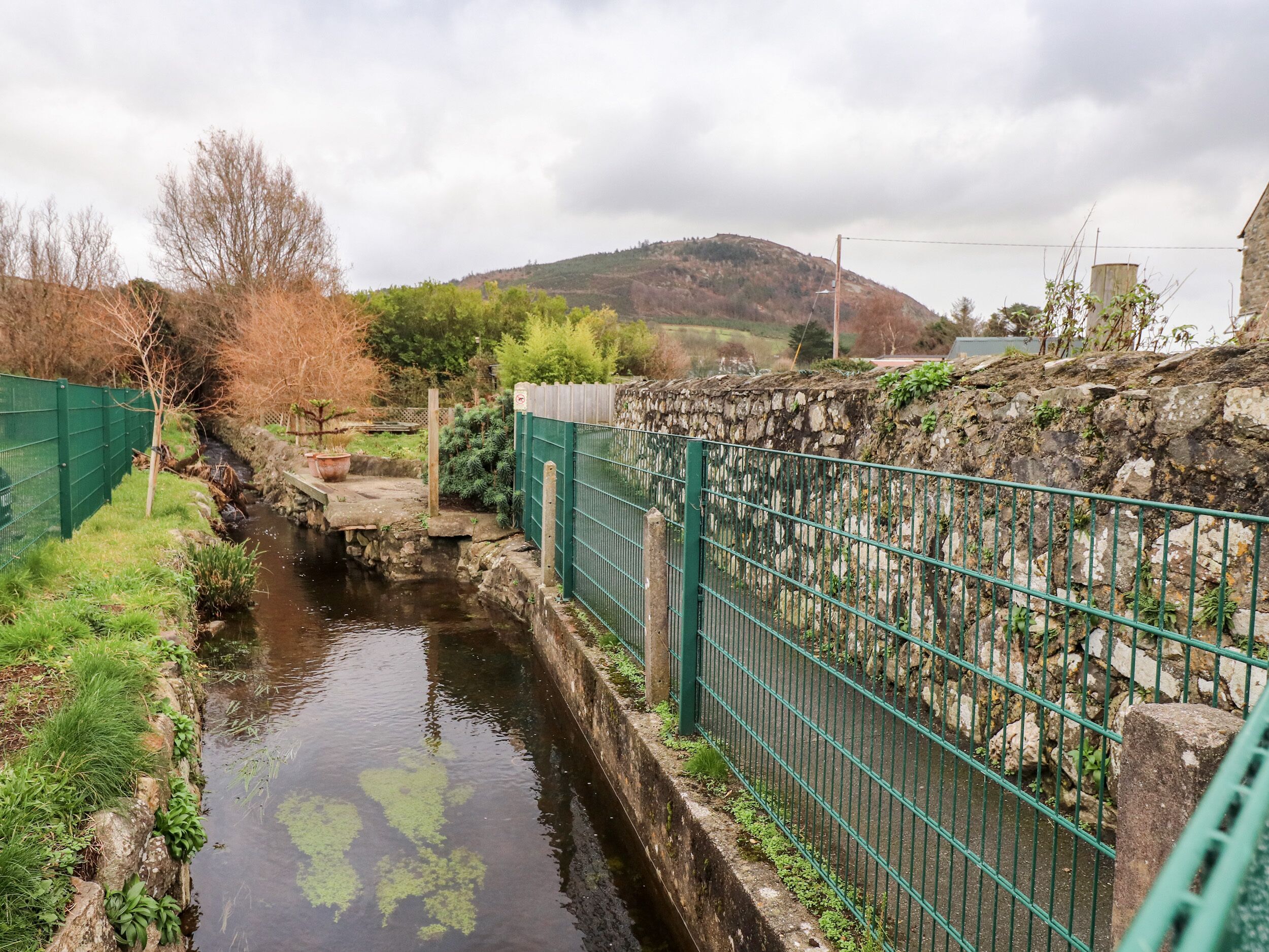 A narrow water channel flanked by stone walls and green fencing, with a stone bridge crossing over it.