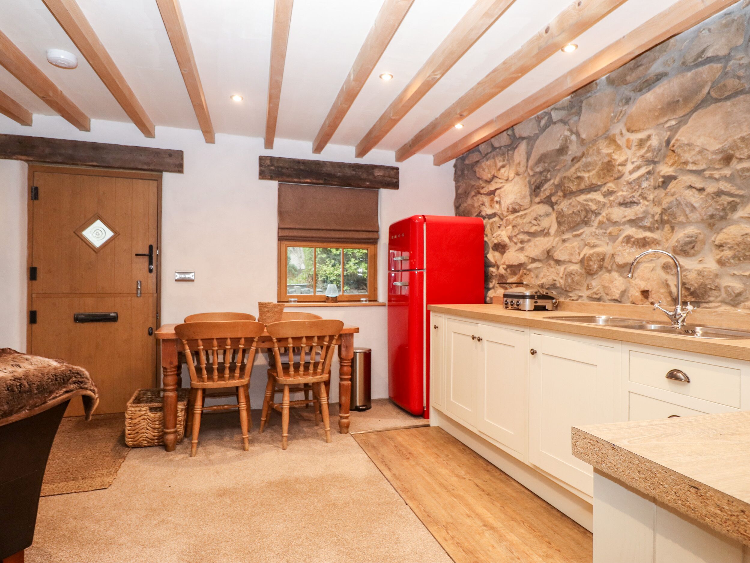 A kitchen with a stone wall, wooden ceiling beams, a red refrigerator, and a dining area with wooden chairs.