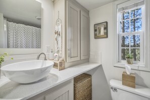A bathroom with a large white sink, a vanity with a mirror, a dreamcatcher, and a window with a view of trees.
