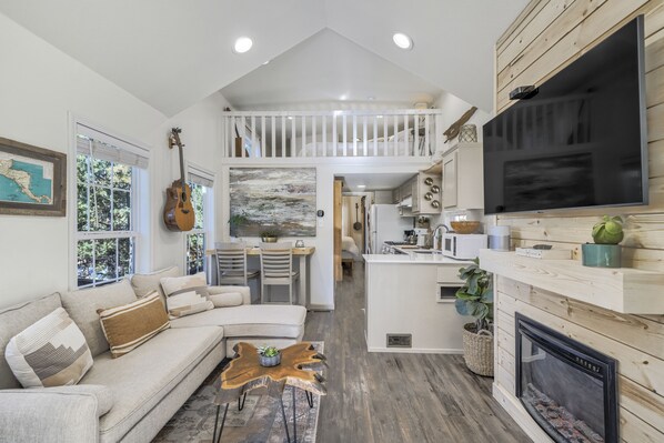 A spacious living area with a high ceiling, a dining table, a kitchen with a large TV, and a fireplace.
