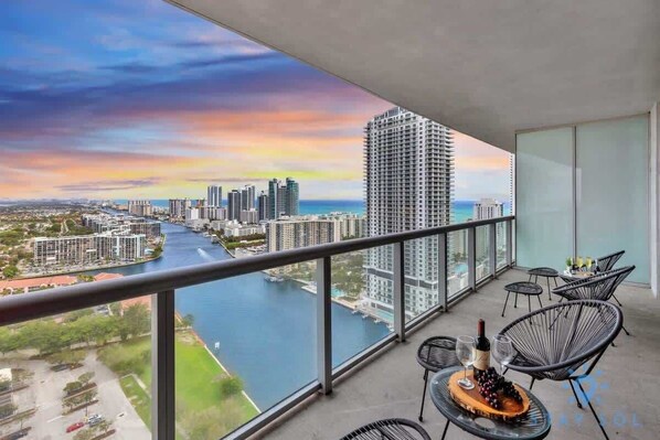 A balcony with a view of a river and cityscape, furnished with chairs and a table with wine glasses and grapes.