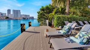 A poolside area with lounge chairs, a view of the city skyline, and a palm tree. - Upper Studio with Stunning Views + Pool + Gym (Hallandale Beach)