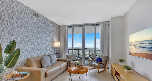 A modern living room with a sofa, armchair, and coffee table, large windows with a city view, and a painting of the ocean.