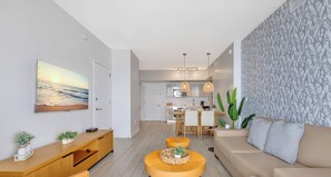 A modern living room with a large painting, wooden furniture, a sofa, and a dining area with a kitchen in the background.