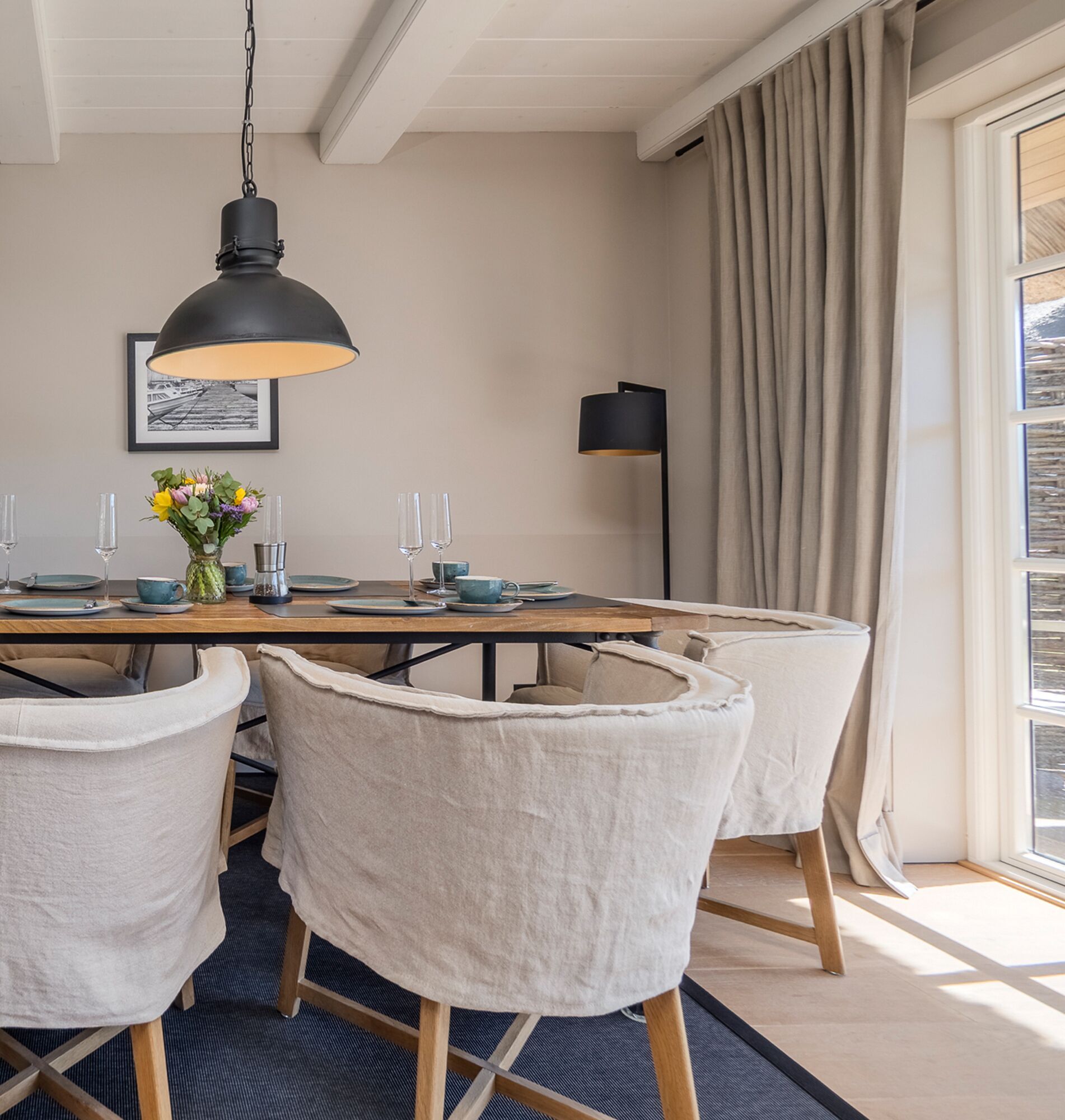 Een eetruimte met een houten tafel, witte stoelen met bekleding, een hangende zwarte hanglamp en een vloerlamp.