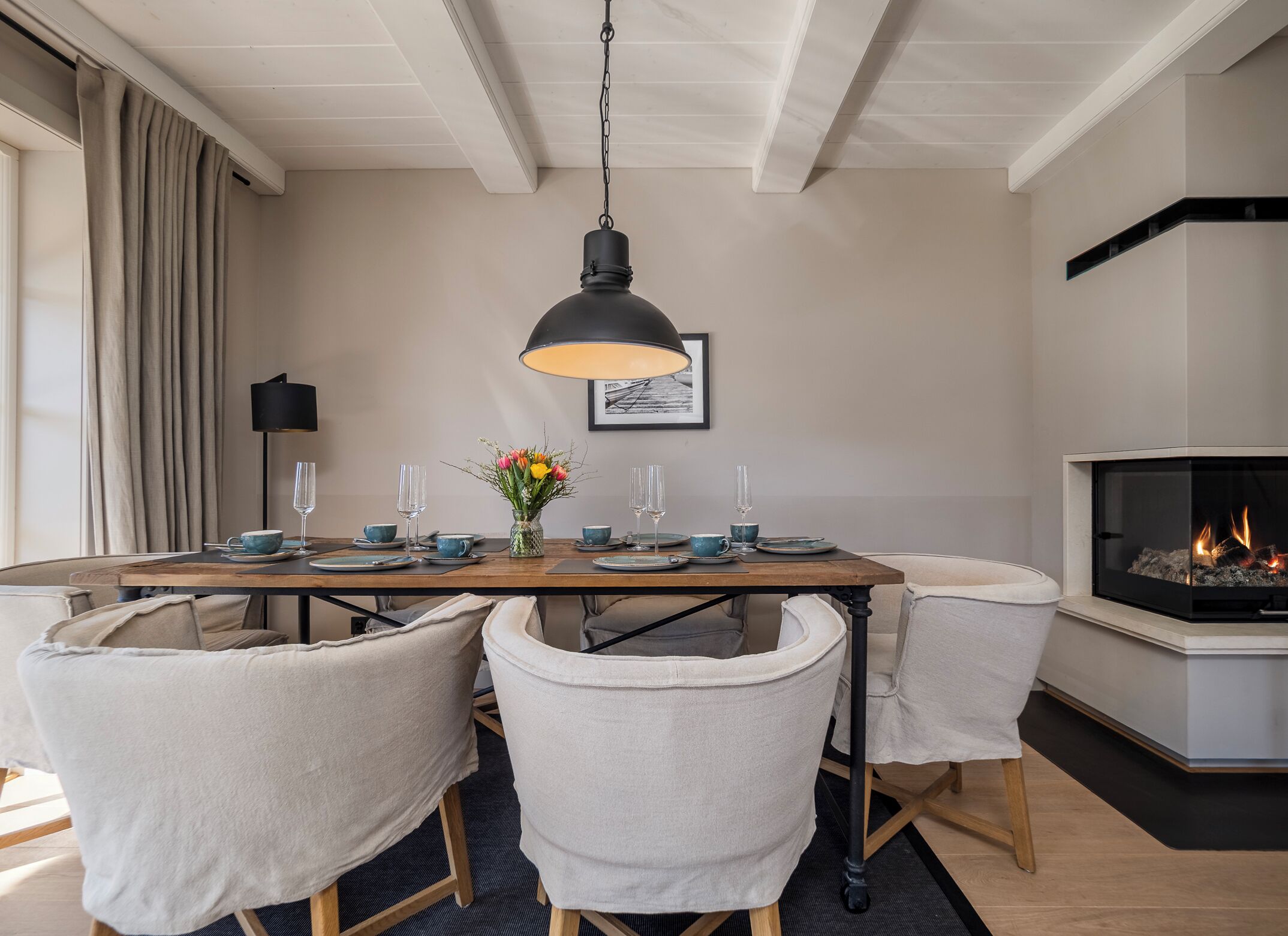 A dining area with a wooden table set for four, surrounded by four chairs, a hanging pendant light, and a fireplace.
