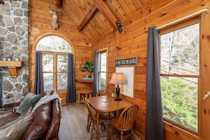 Ein gemütliches Blockhäuschen mit Kamin aus Stein, Holzwänden, einem Esstisch mit Stühlen und großen Fenstern mit Blick auf den Wald.