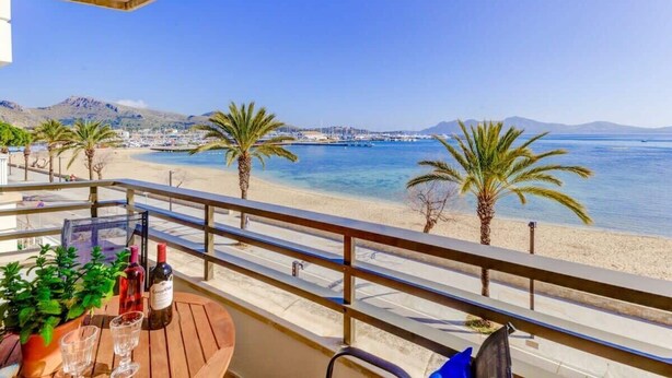 Un balcón con una mesa lista para dos personas, con vista a una playa con palmeras y un mar azul cristalino.