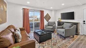 A modern living room with a brown leather sofa, patterned armchairs, a black ottoman, and a patterned rug. A flat-screen TV is mounted on the wall, and a large window with curtains is visible. - 6A Family Friendly RedCliff Condo POOL & HOT TUB (Moab)