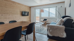 Ein modernes Wohnzimmer mit einer Holzwand, einem Esstisch mit Stühlen, einem Sofa mit Felldecke, einer Stehlampe und einem an der Wand montierten Fernseher.