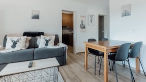 Ein modernes Wohnzimmer mit einem grauen Sofa, einem Holzespeisetisch mit vier Stühlen und einer durch einen offenen Durchgang einsehbaren Küche.
