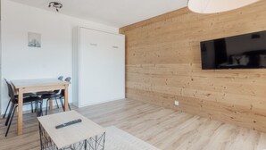 Ein modernes Zimmer mit einer Holzwand, einem Flachbildfernseher, einem Esstisch mit Stühlen und einer Fernbedienung auf einem kleinen Tisch.