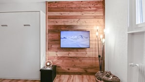 Ein Zimmer mit Holzpaneelen, einem montierten Fernseher, einem schwarzen Ottomanen und einem Korbwäschekorb.