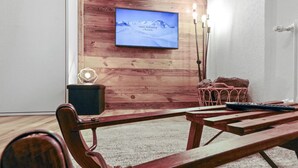 Ein gemütliches Zimmer mit einer Holzwand, einem angebrachten Fernseher, einer Holzbank, einem kleinen Tisch und einem pinken Stuhl.