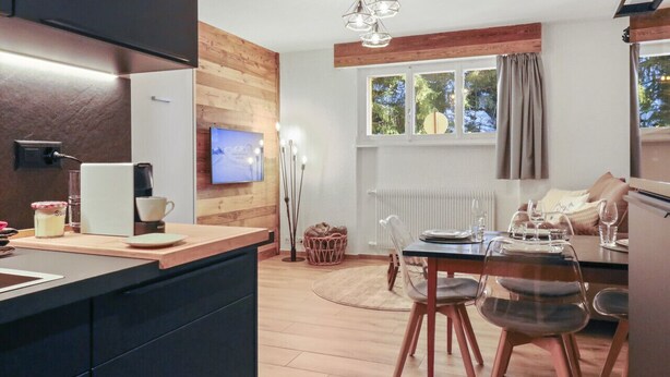 Eine moderne Küche mit einer Wand in Holzoptik, ein Essbereich mit Tisch und Stühlen sowie ein großes Fenster mit Vorhängen.