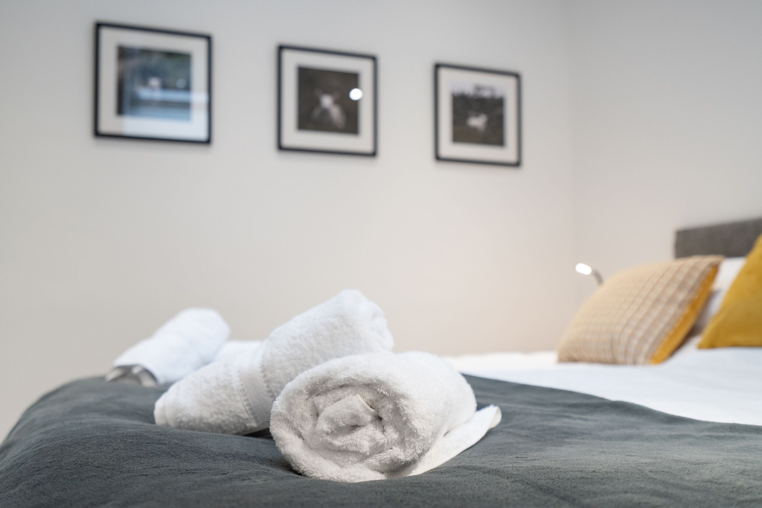 A neatly made bed with rolled white towels, a gray blanket, and a yellow pillow. Three framed pictures are hung on the wall above the bed.