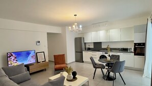 Un salon moderne avec un canapé, une table basse et un téléviseur. On y trouve également un coin repas avec des chaises et une table. La cuisine est visible et se distingue par ses armoires et ses appareils électroménagers blancs.