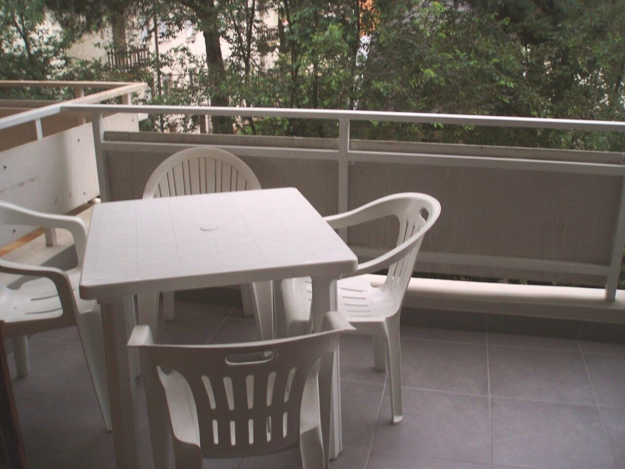 Un balcon avec une table et des chaises blanches, une balustrade et une vue sur les arbres.