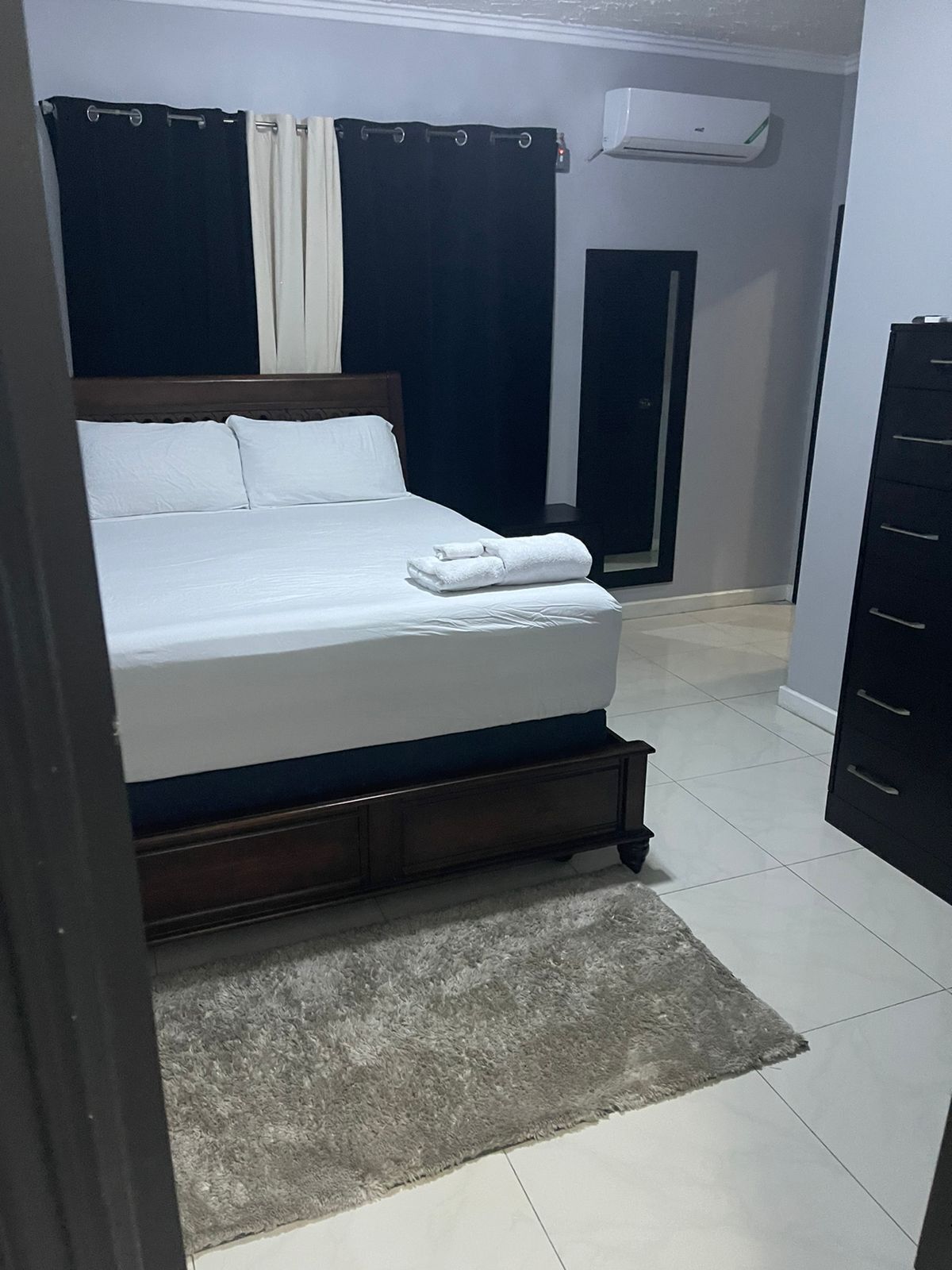 A bedroom with a bed, a dark-colored dresser, a white air conditioning unit on the wall, and a door.