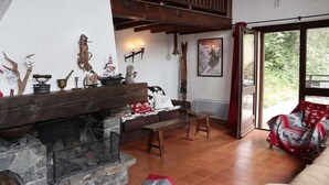 A cozy living room with a stone fireplace, wooden furniture, and a large window offering a view of greenery. - Chalet for 11, slopeside, fireplace, Les Orres (Les Orres)