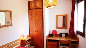 A bedroom with a bed, wooden wardrobe, desk with chair, and a hanging lamp.