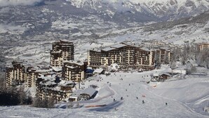 A snow-covered ski resort with multi-story buildings, ski slopes, and a mountainous backdrop.
