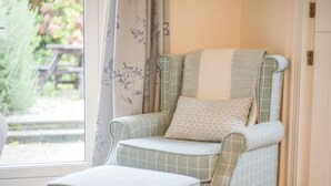 A cozy room with a plaid armchair and ottoman, a window with floral curtains, and a wooden floor. - The Old Lace Shop (Seaton)