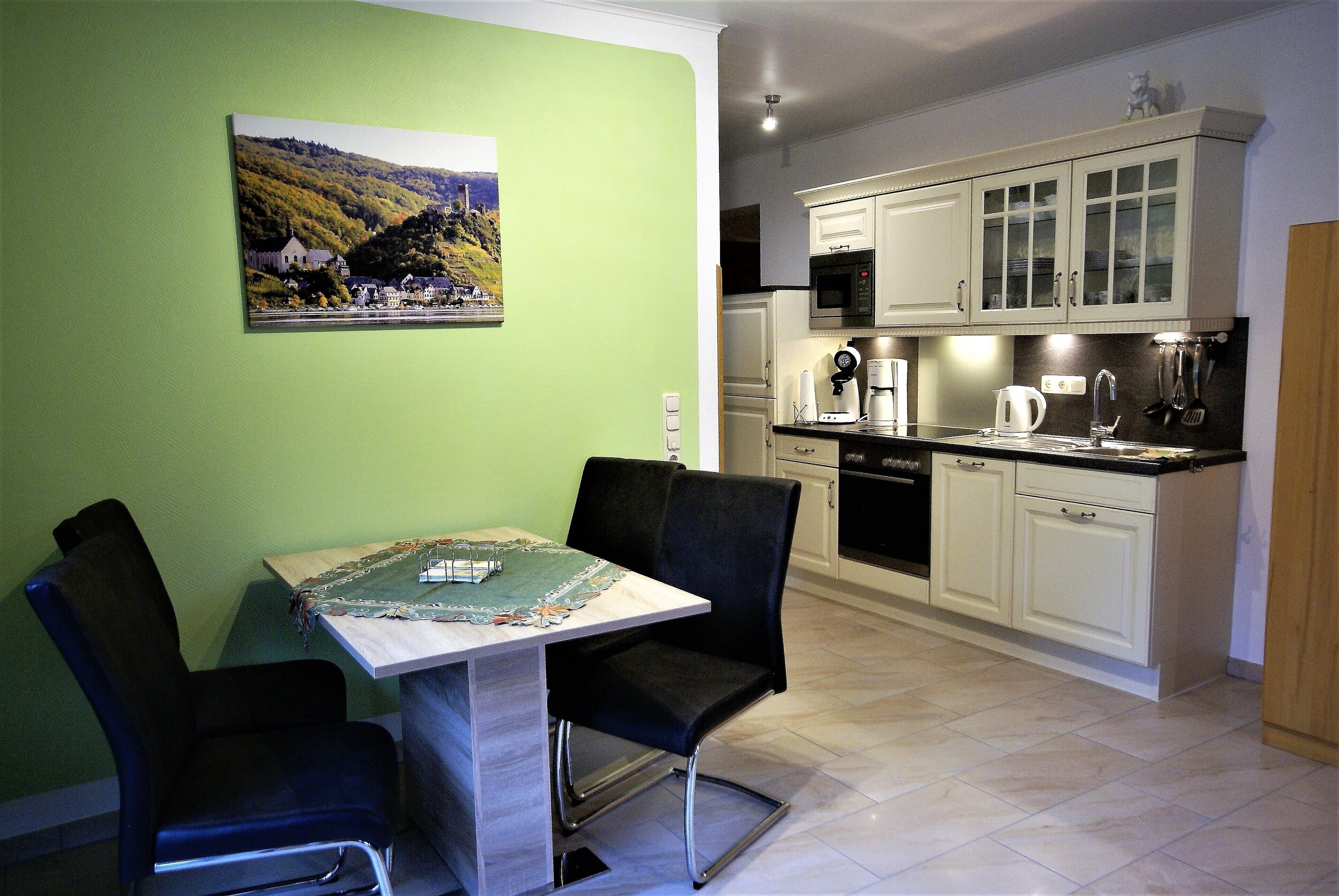 A dining area with a table and chairs, a kitchen with white cabinets and appliances, and a green wall with a landscape painting.