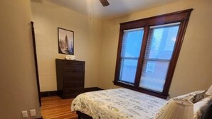 Un dormitorio con una cama, un escritorio con lámpara, un ventilador de techo, una ventana con persianas y un aparador.