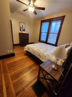 A bedroom with a bed, a desk with a lamp, a ceiling fan, a window with blinds, and a dresser.