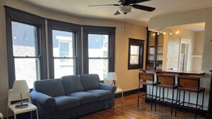 Una habitación con un sofá azul oscuro, una mesita blanca pequeña, una zona de bar con taburetes y ventanas grandes.