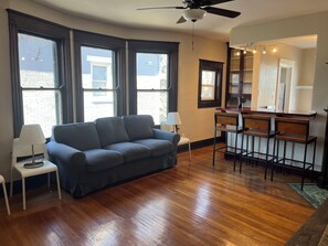 A room with a dark blue sofa, a small white table, a bar area with stools, and large windows.