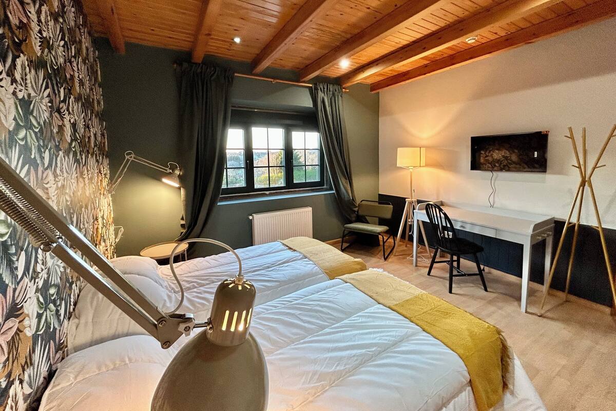 A bedroom with two beds, a desk, a chair, a lamp, a radiator, and a window with curtains.