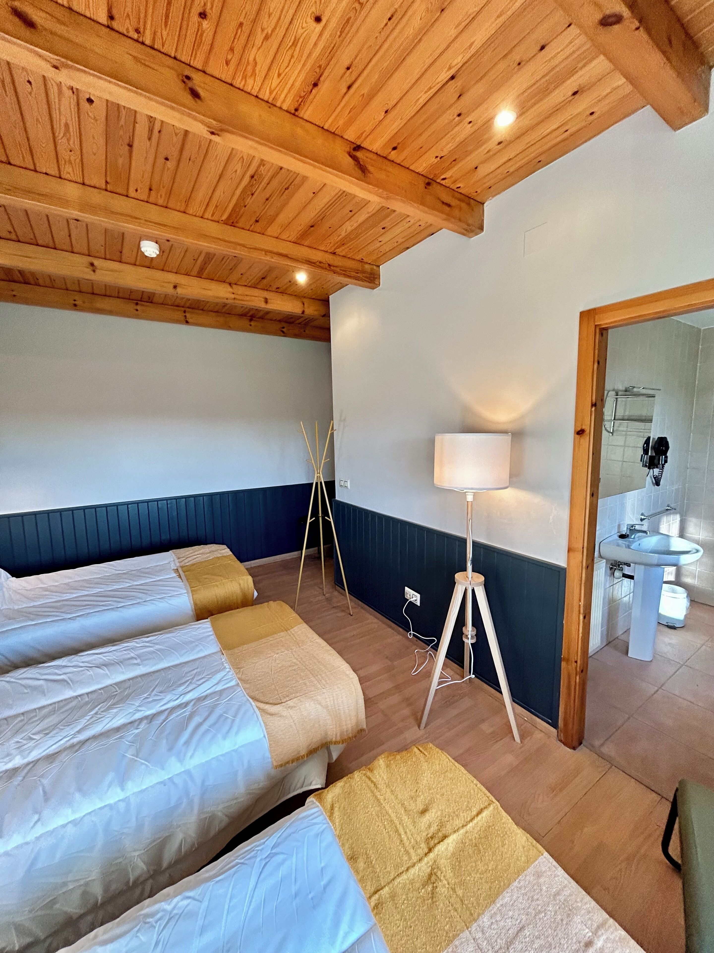 A room with wooden ceiling beams, a bed with white and beige bedding, a floor lamp, and a bathroom visible through an open door.