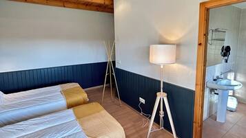 Quarto com vigas de madeira no teto, cama com roupa de cama branca e bege, luminária de chão e banheiro visível por uma porta aberta.
