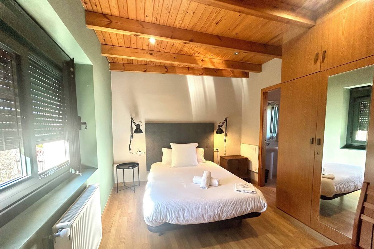 A bedroom with a large bed, wooden ceiling beams, a radiator, a nightstand, a mirror, and a window with blinds.