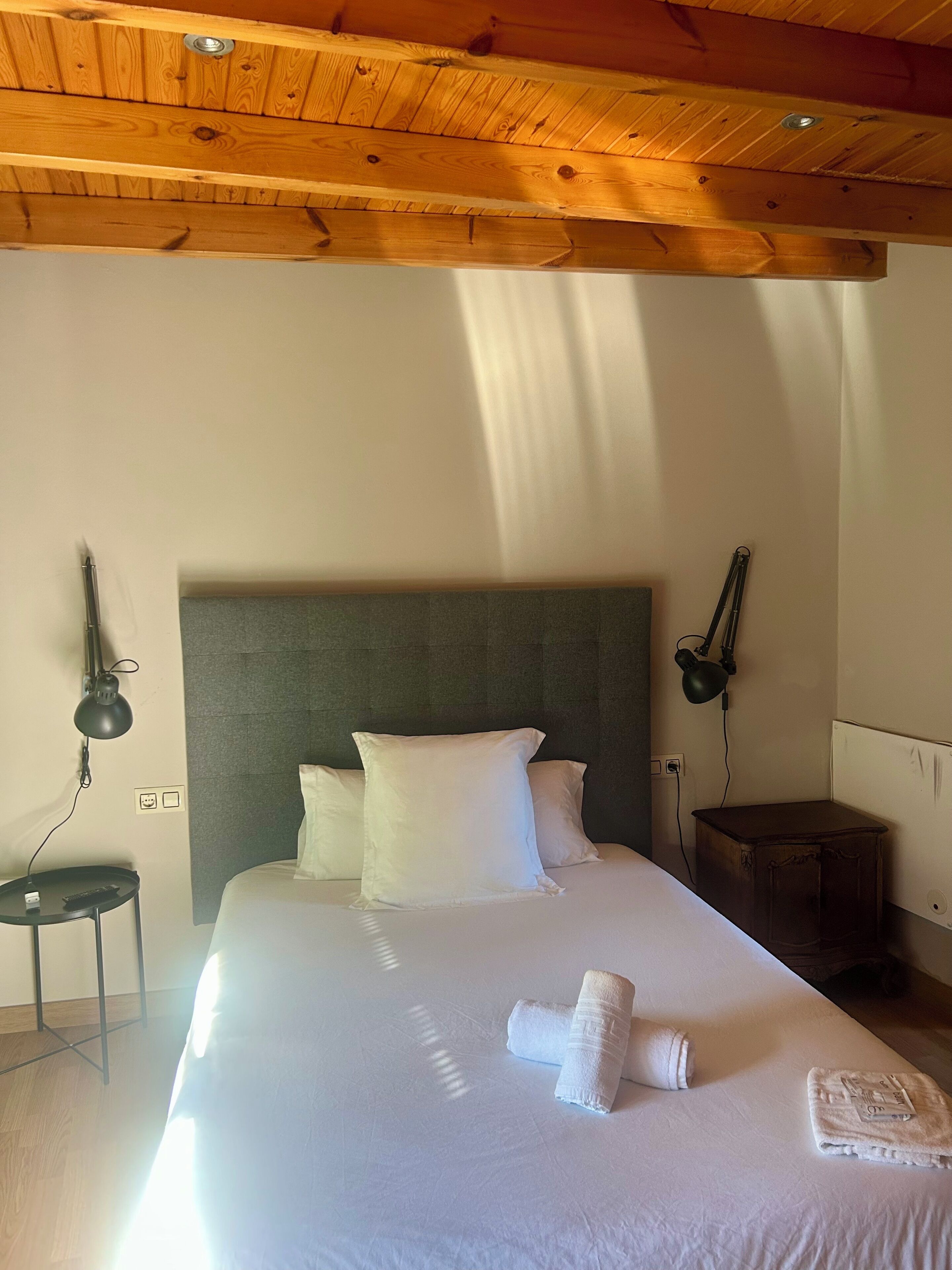 A hotel room with a wooden ceiling, a bed with white linens, a headboard, two bedside lamps, a small table, and a rolled towel on the bed.