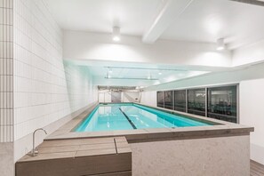 A modern indoor swimming pool with clear glass walls, a tiled floor, and a wooden deck area.