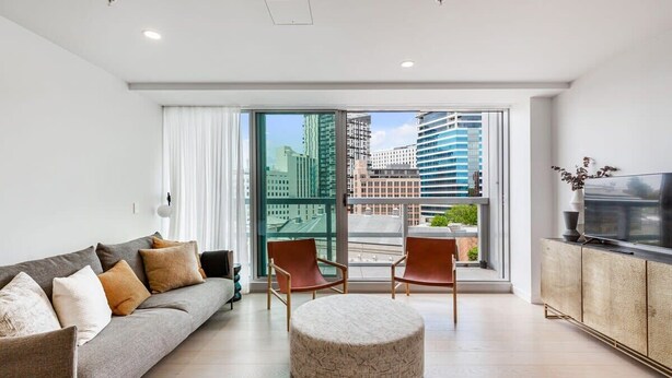 Un salon moderne comprenant un canapé, un pouf, des fauteuils, un téléviseur et un balcon offrant une vue sur la ville.