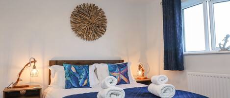 A neatly made bed with blue bedding and decorative pillows, a bedside table with a lamp, and a wall art piece.