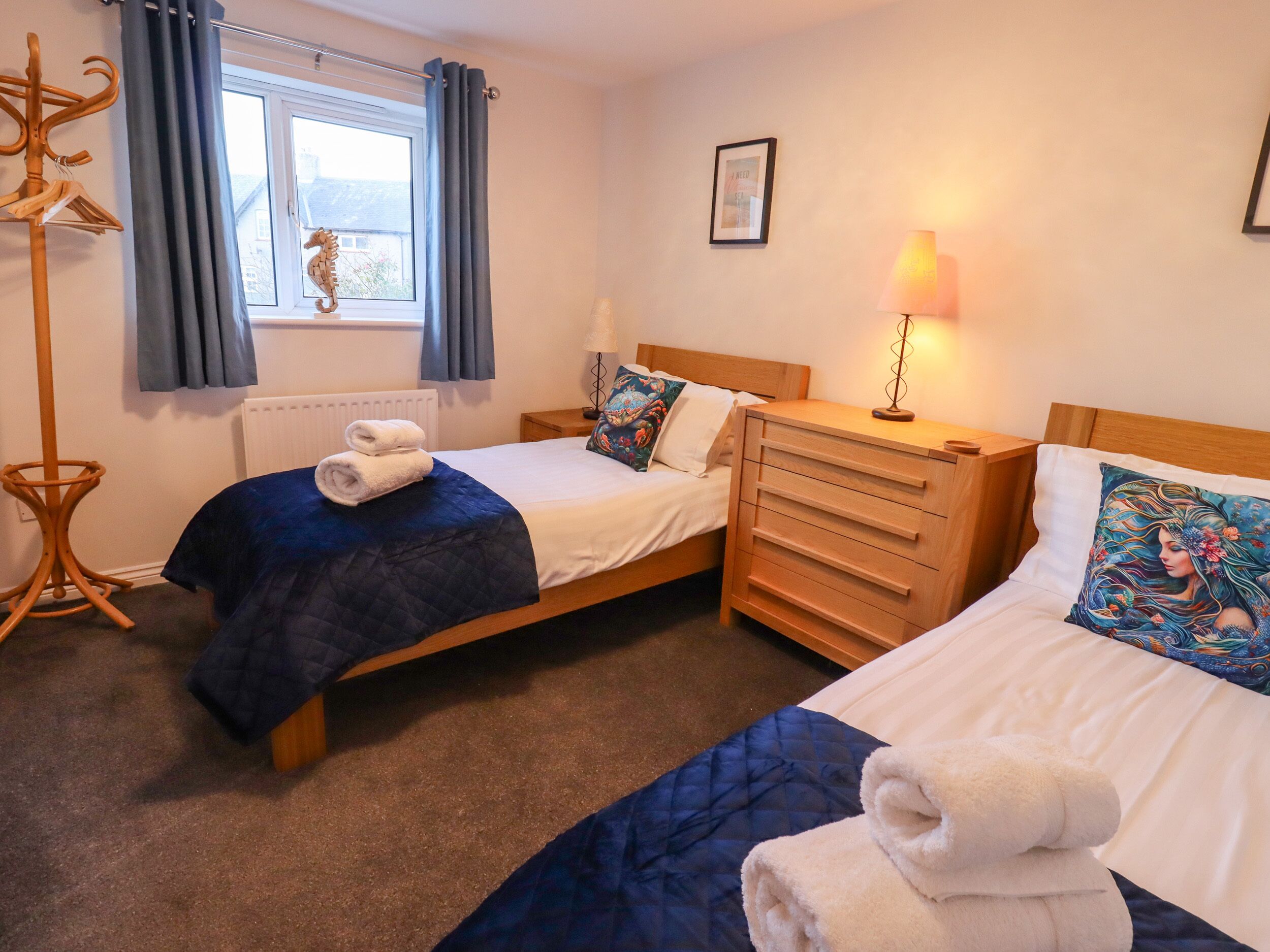 A hotel room with two beds, a wooden dresser, a floor lamp, a coat rack, and a window with curtains.