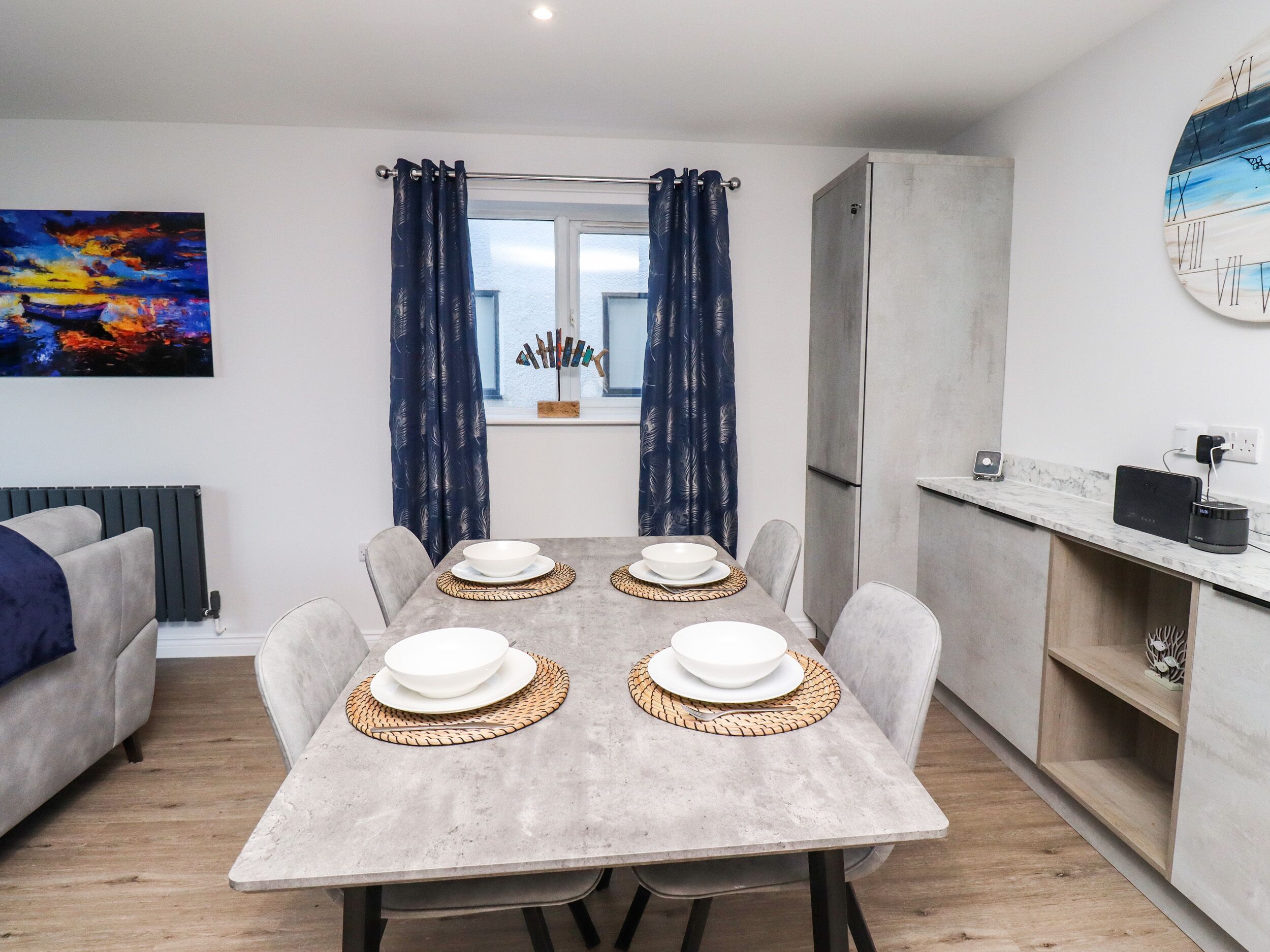 A modern dining area with a grey table set for four, a grey sofa, a window with blue curtains, and a wall clock.
