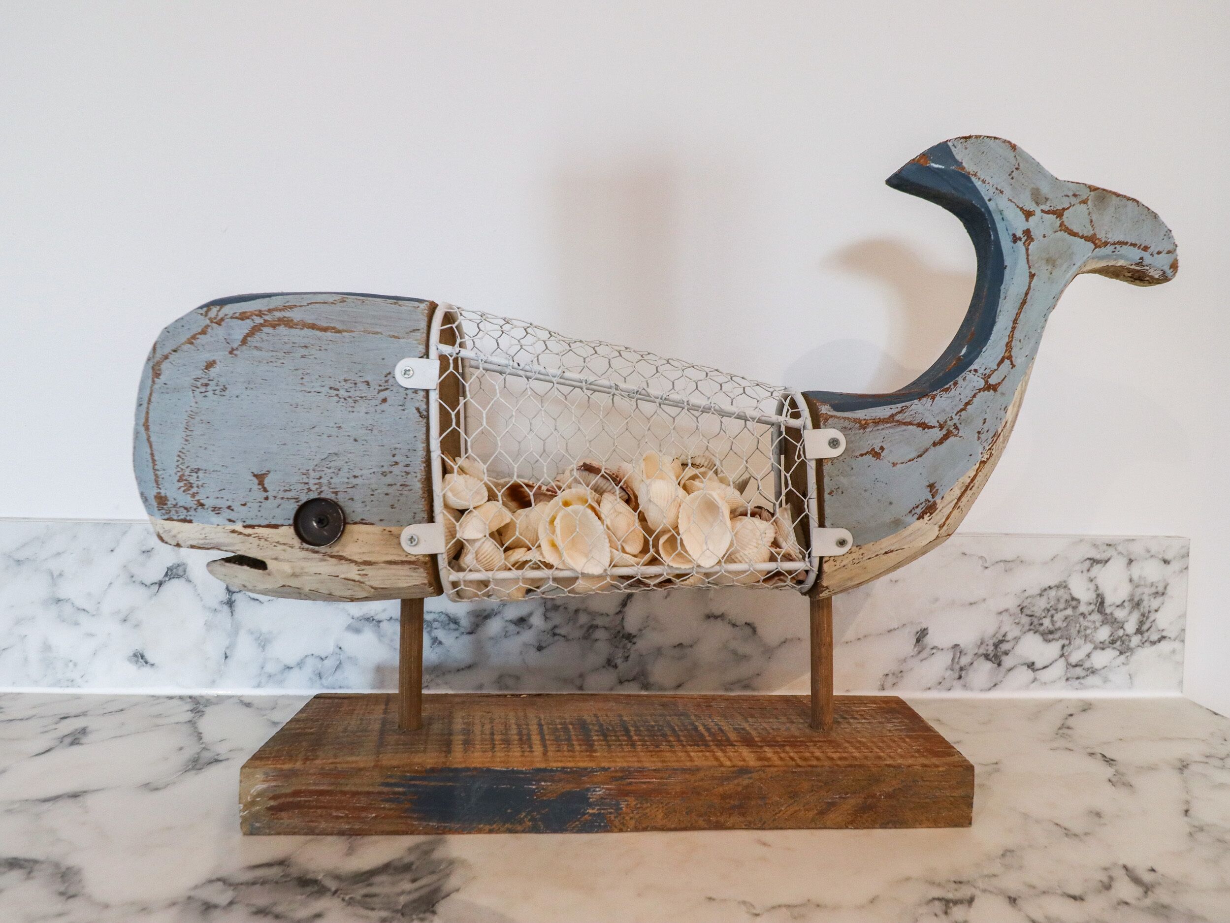A rustic whale sculpture with a wire basket filled with seashells, mounted on a wooden base.
