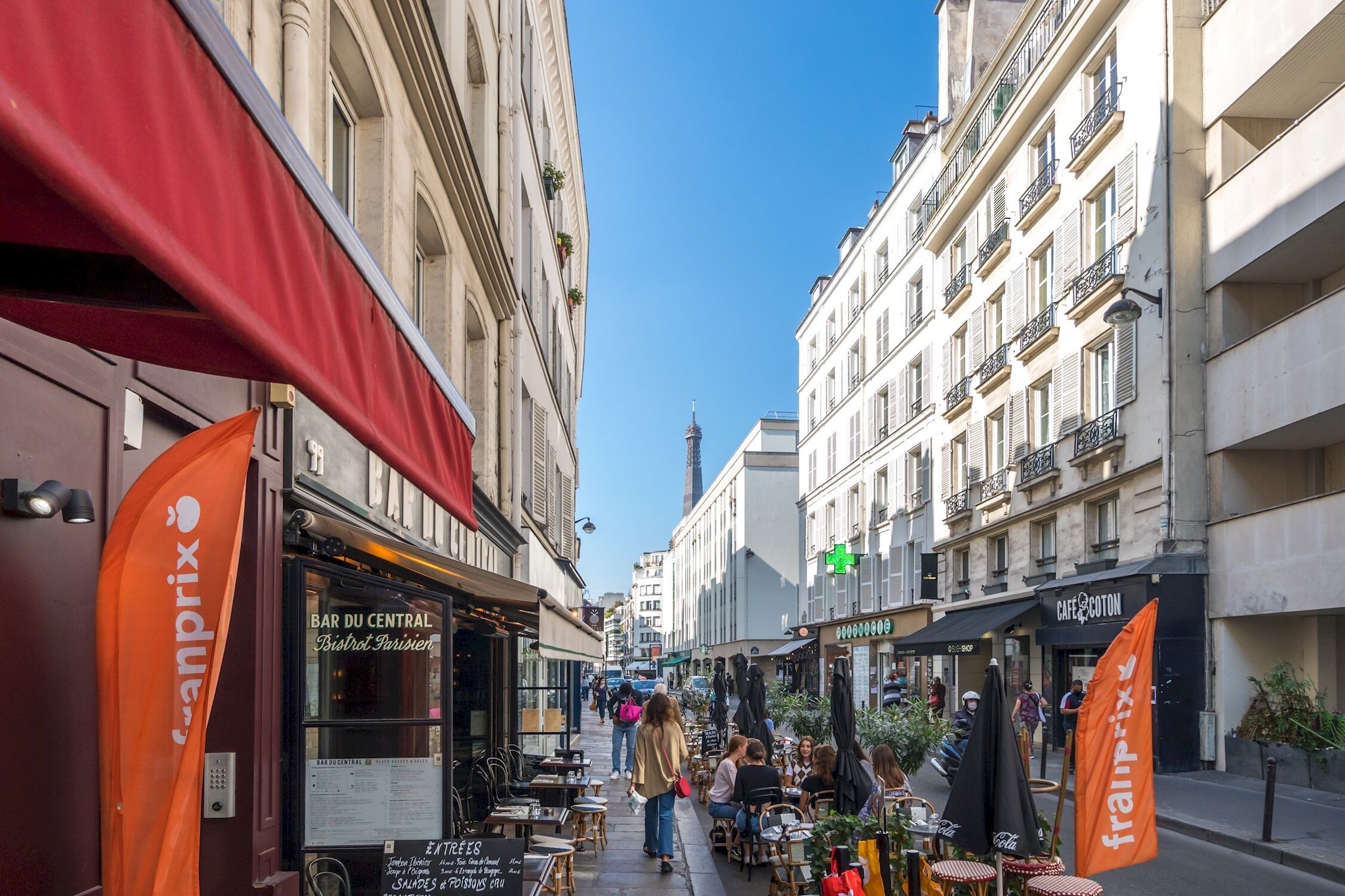Une rue étroite bordée de cafés avec des terrasses, une marquise rouge bien visible et, au fond, un immeuble imposant.