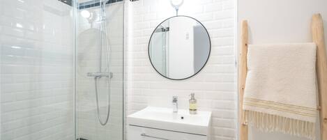 A bathroom with a glass-enclosed shower, a white sink with a round mirror, and a wooden towel rack.