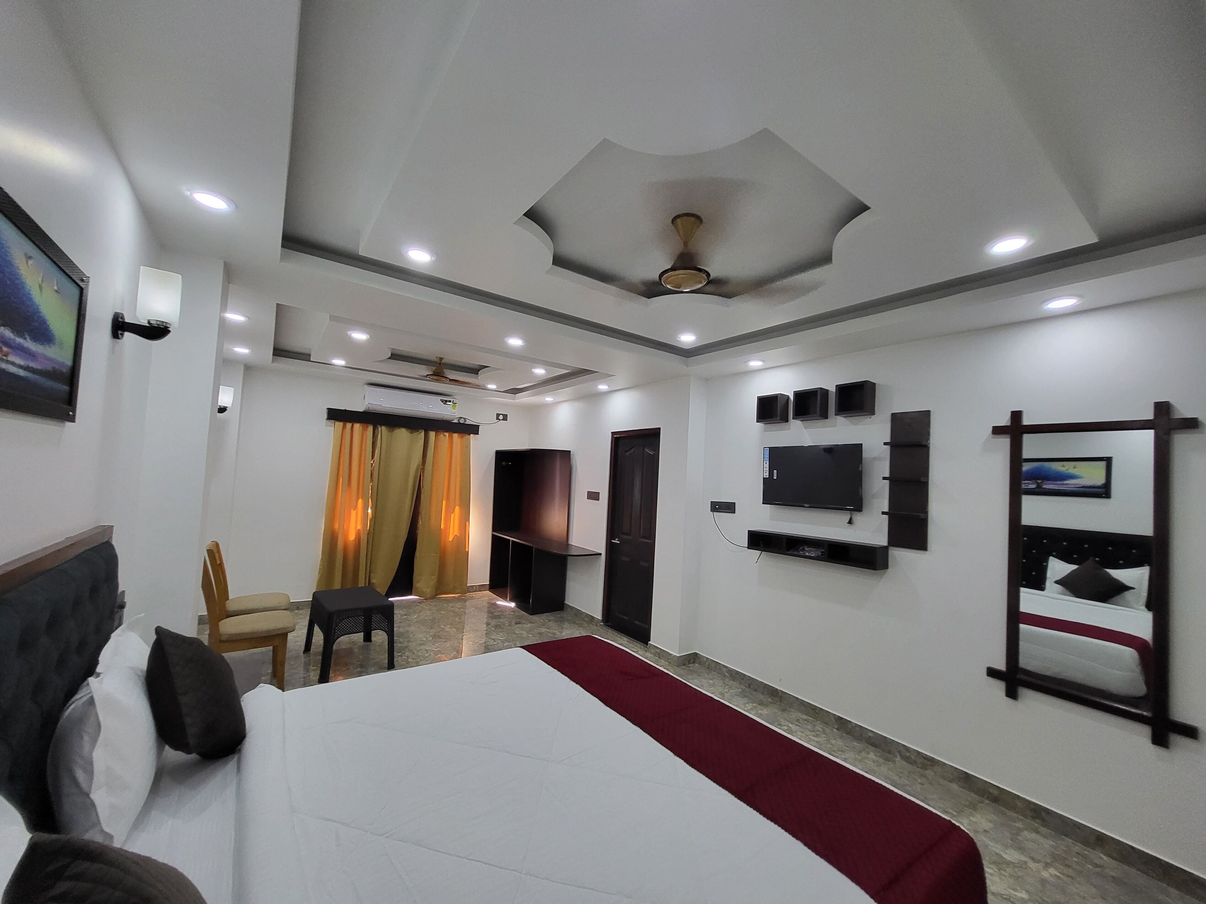 A hotel room with a bed, a TV mounted on the wall, a ceiling fan, and a window with curtains.